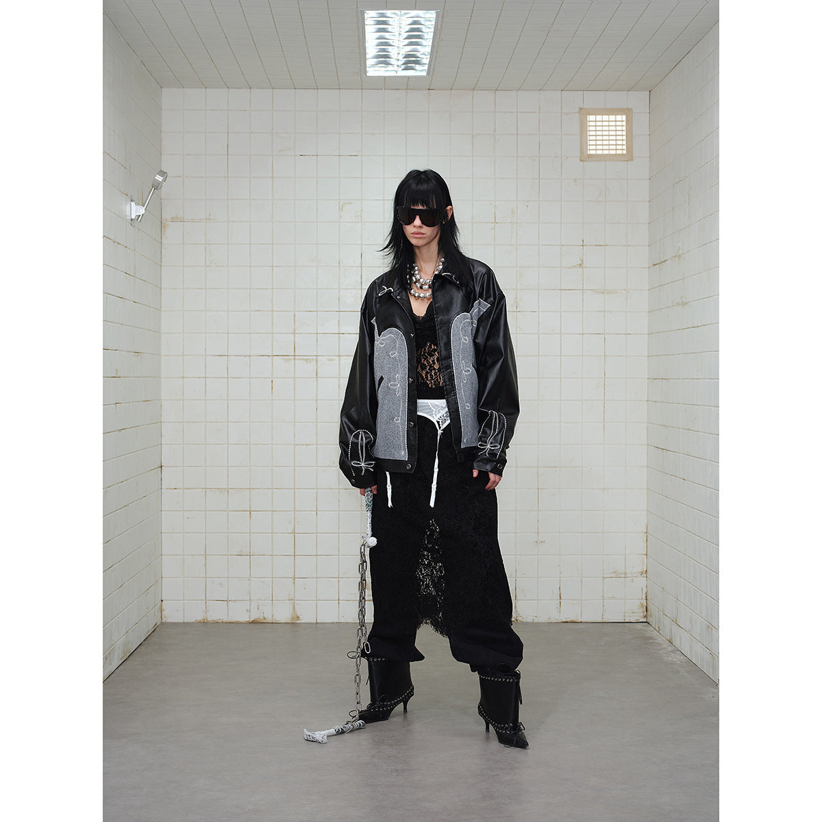Model wearing a black jacket with gray embroidered panels and white stitching, styled with a black lace skirt and boots.