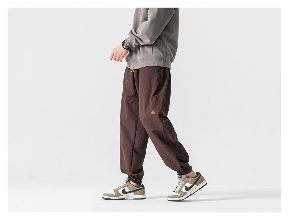 Side view of a model wearing loose dark brown jogger pants with elastic cuffs and a small orange leg emblem.