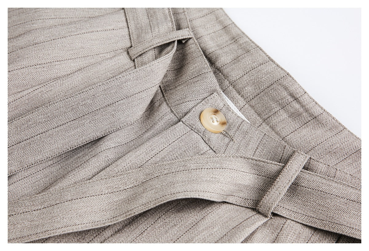Close-up of taupe-gray herringbone trousers showing a tan waistband button, belt loops, and front closure.