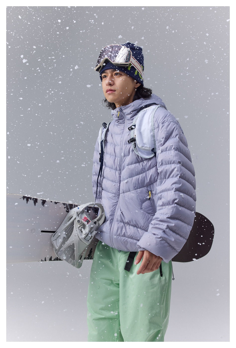 Person in a light purple hooded puffer jacket with a snowboard, goggles, beanie and green pants in falling snow