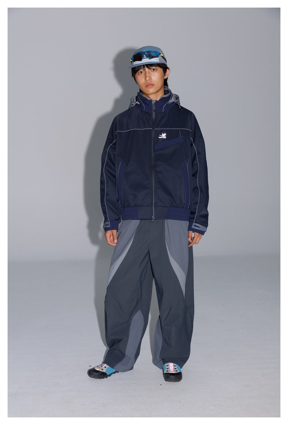 Model wearing a dark navy hooded jacket and loose gray panelled pants, shown full length against a gray studio backdrop.