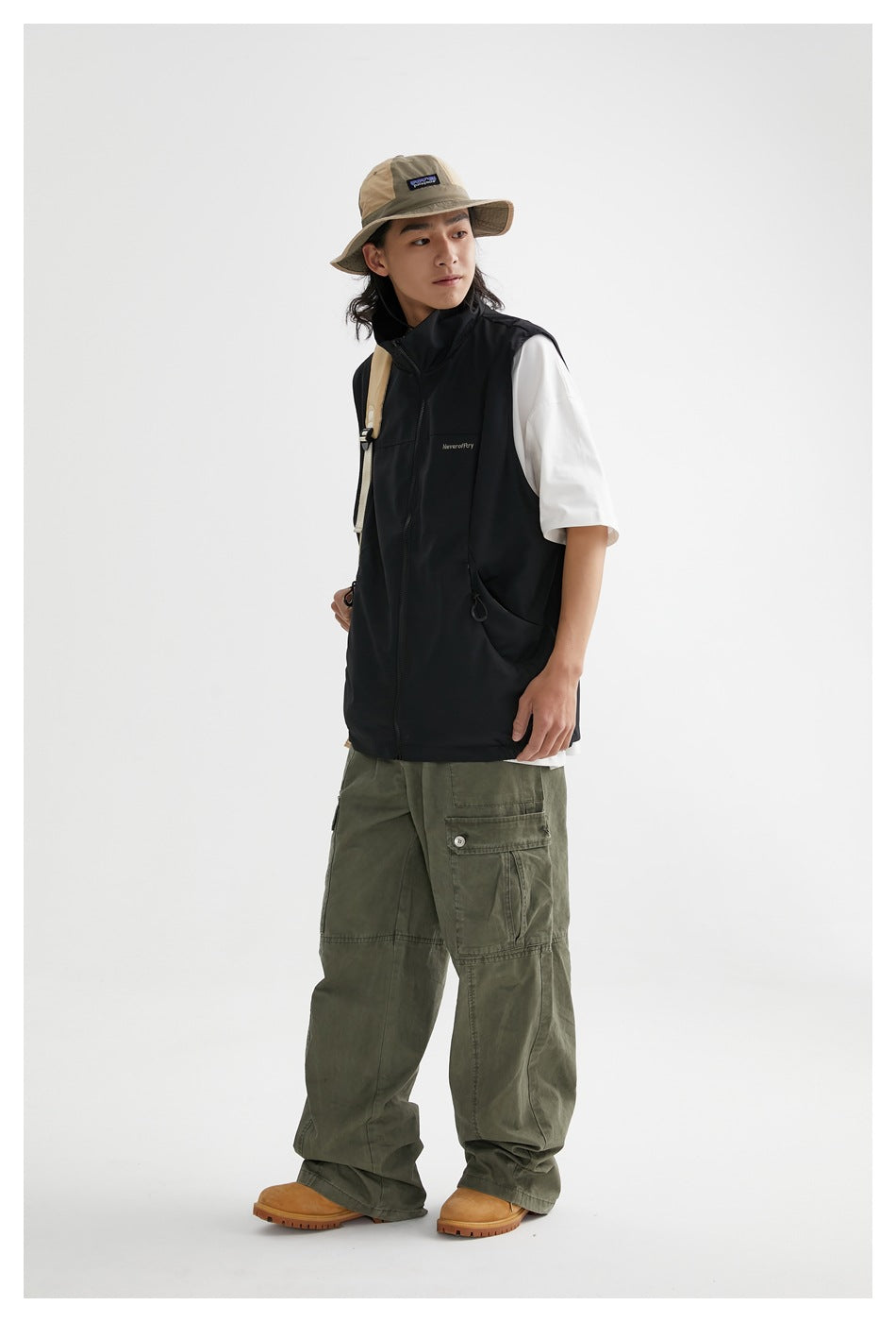 Full-length model wearing a black zip-front vest over a white T-shirt, olive cargo pants, and a tan wide-brim hat.