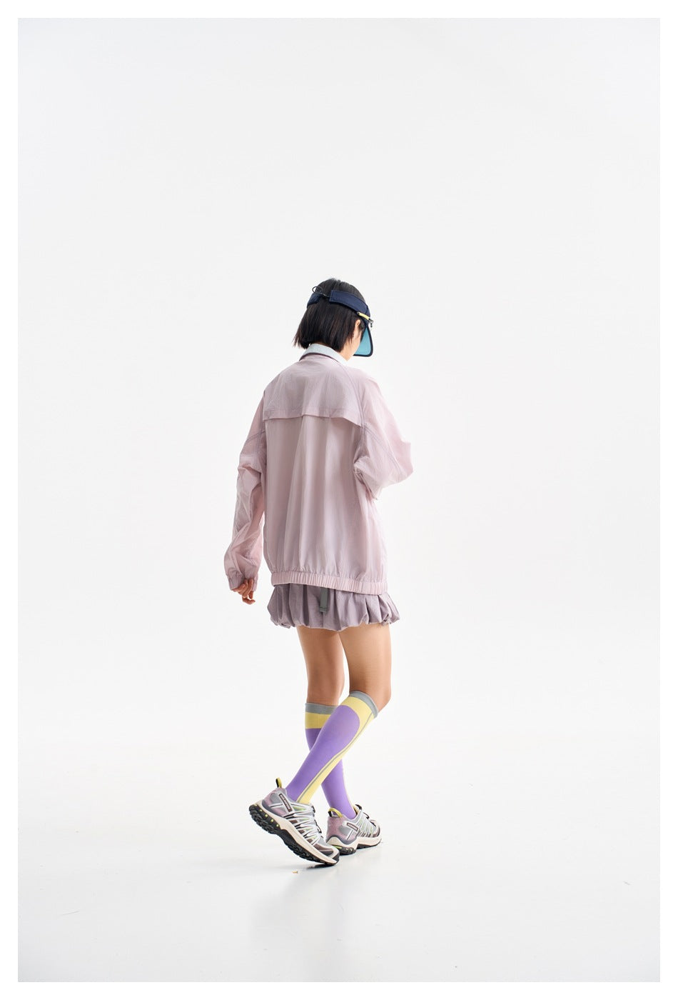 Rear view of a model wearing a pale pink jacket with a back yoke and elastic hem, paired with a pleated skirt.