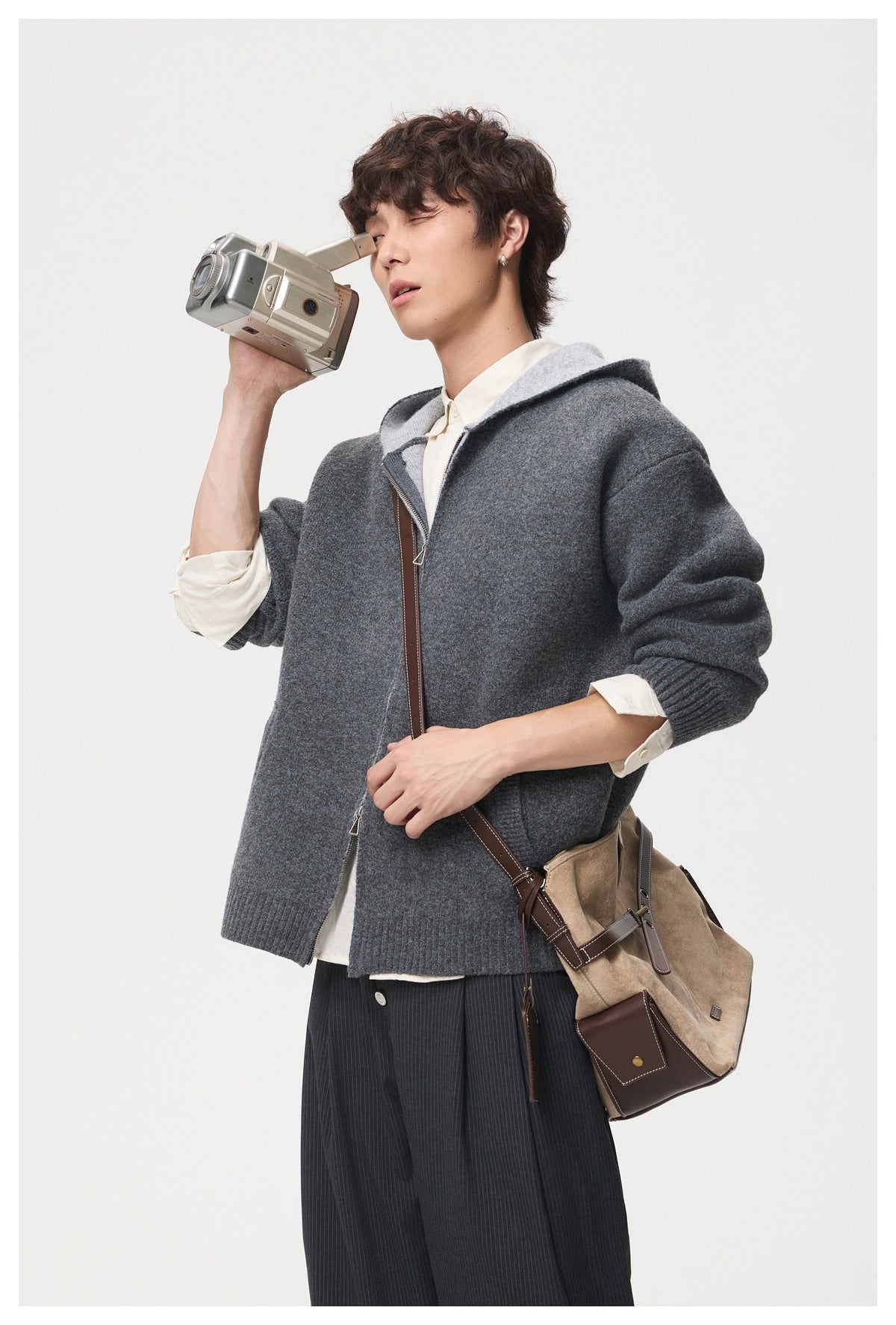 Model wearing a charcoal hooded cardigan while holding a camcorder and carrying a tan shoulder bag.