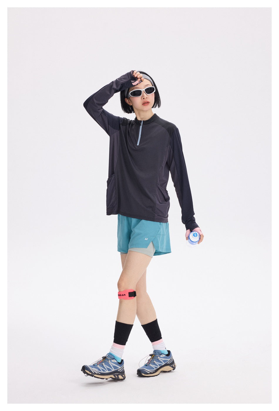 A woman wears a dark charcoal long-sleeve quarter-zip top with blue trim and turquoise running shorts.