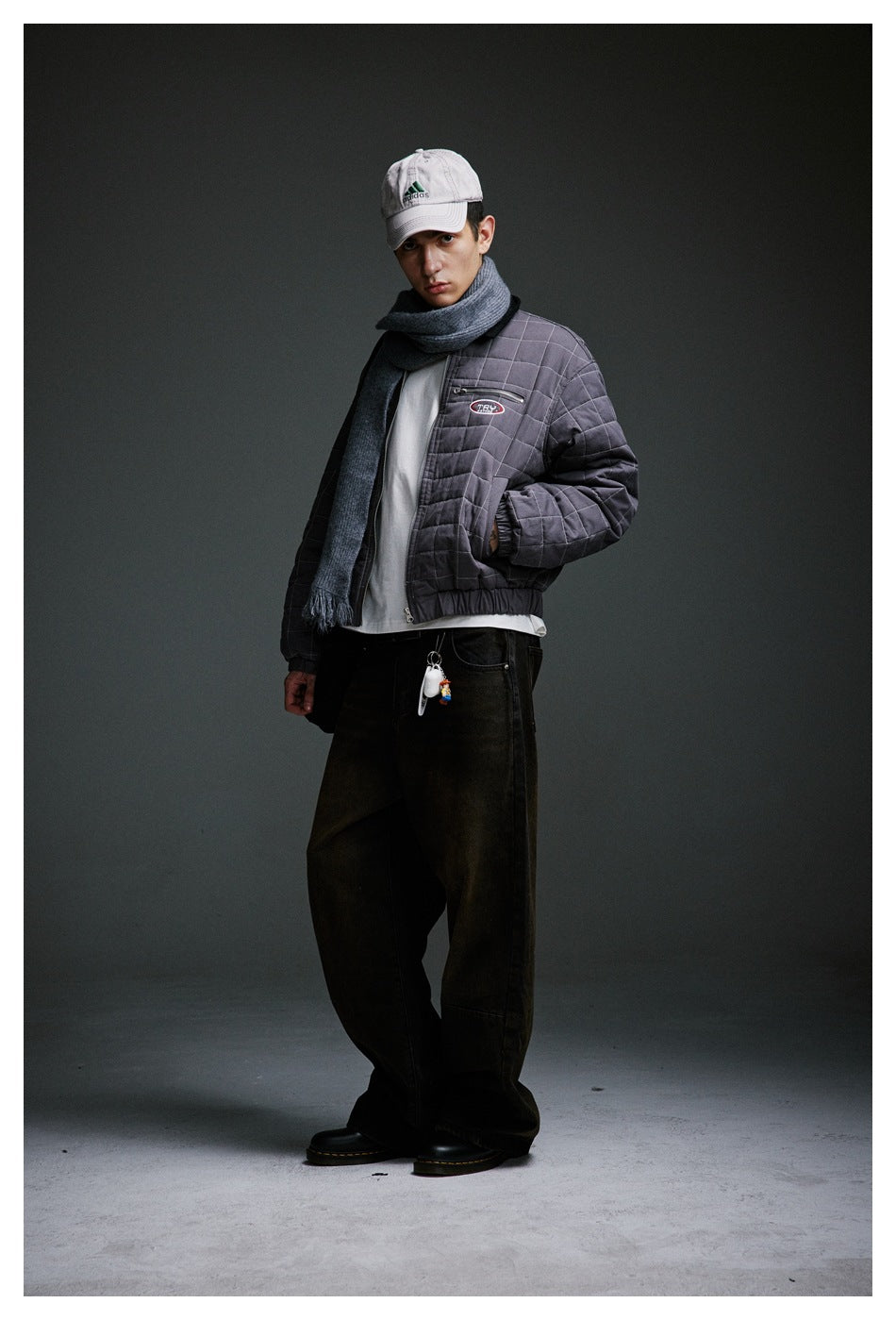 Model in a gray quilted jacket and scarf, shown at an angle with dark wide-leg trousers and cap.