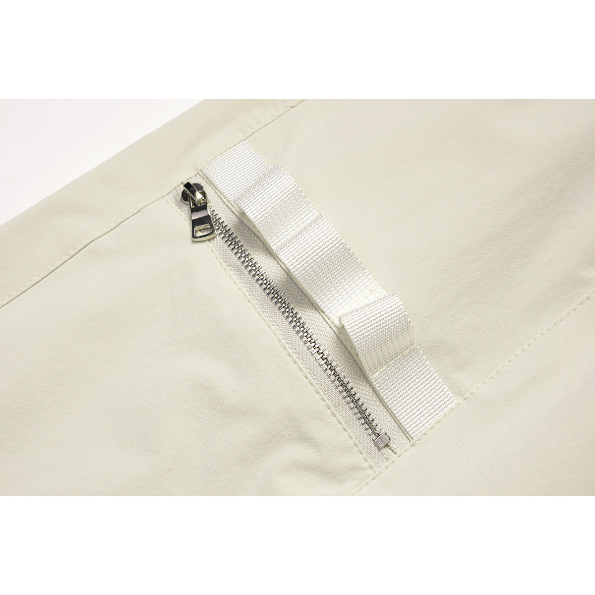 Close-up of an off-white pants pocket with a metal zipper and woven fabric tab.