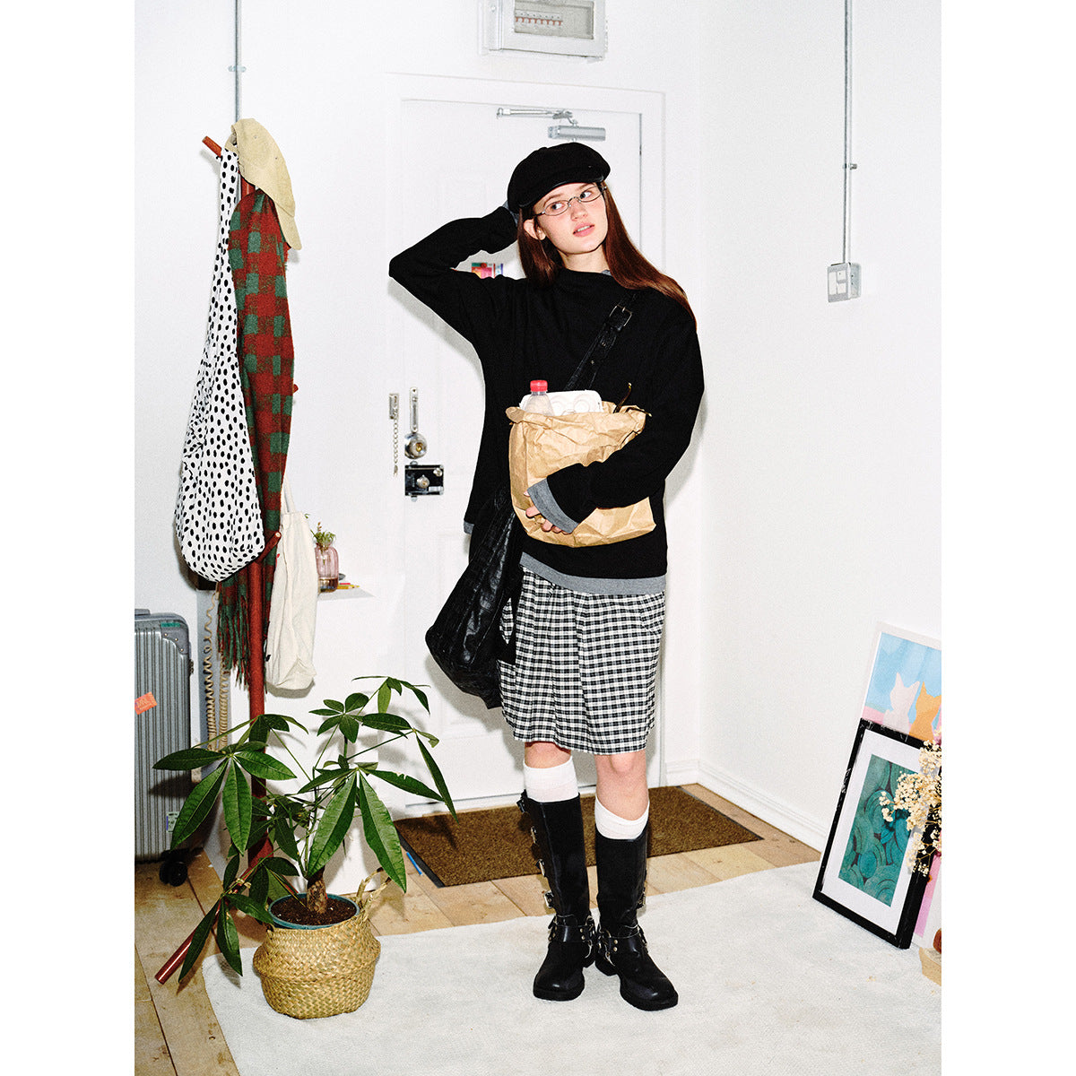 Model in a black sweater with gray trim, checked skirt, cap and tall boots, holding a paper bag.