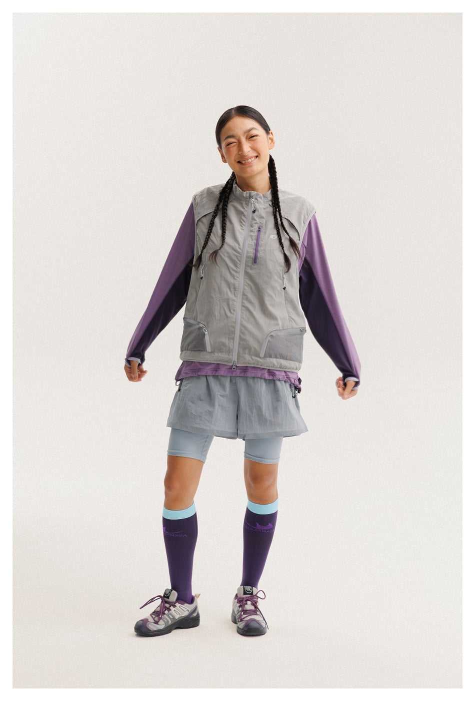 Full-length front view of a model wearing a gray zip vest, purple long-sleeve top, gray shorts, and purple socks.