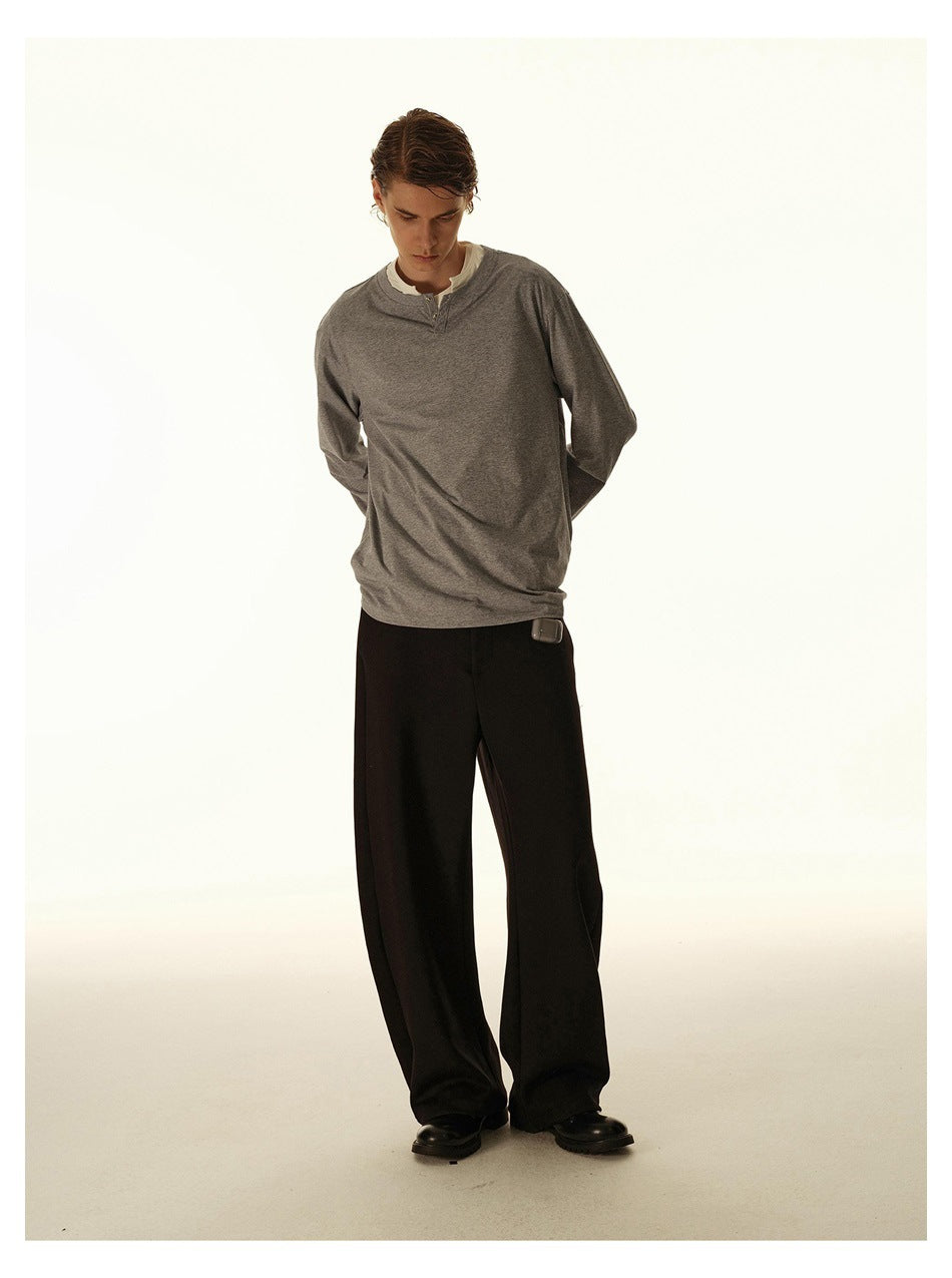 Model wearing a gray long-sleeve henley over a white shirt and wide black trousers, standing with hands behind his back.