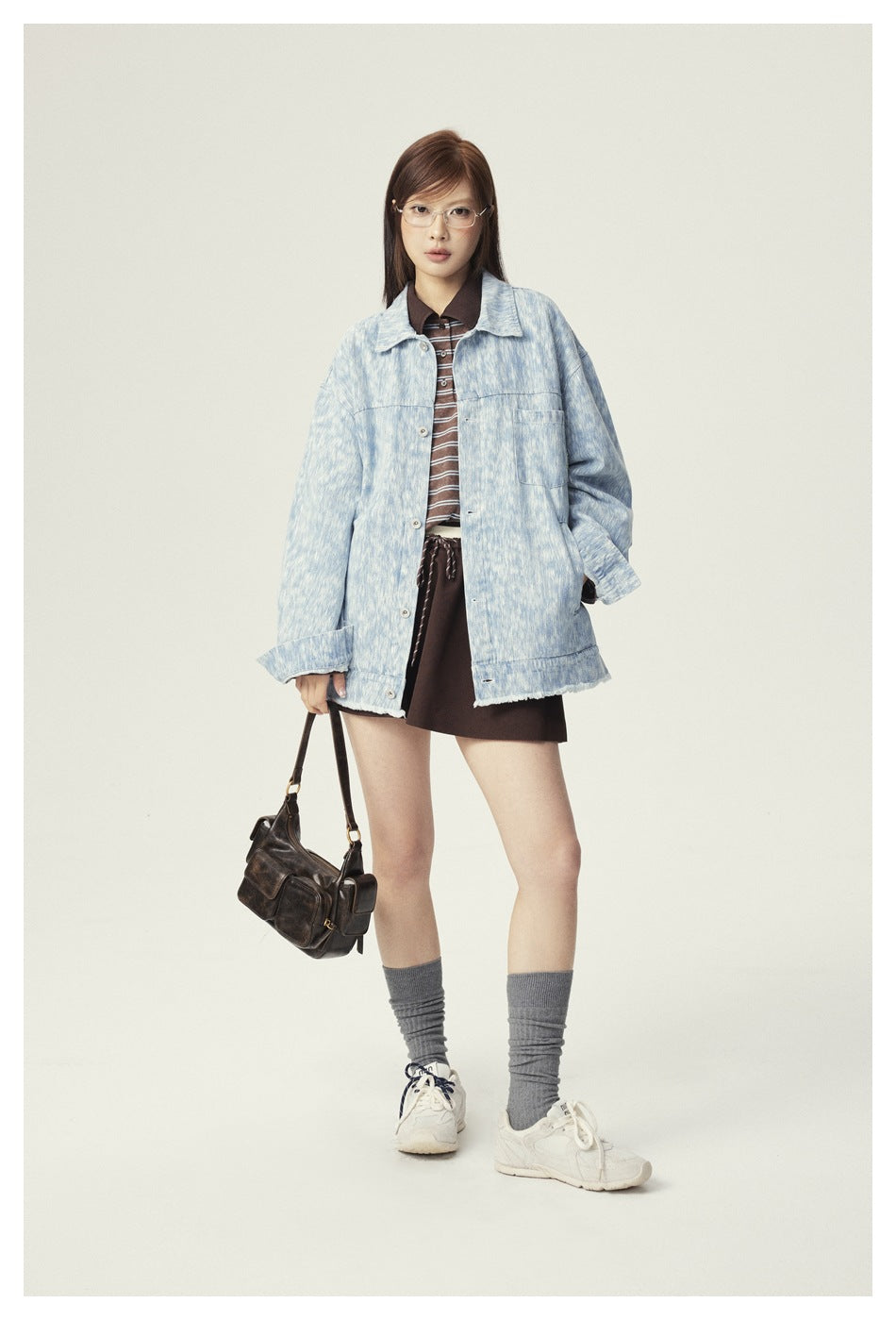 Model wearing a light blue washed denim jacket open over a striped polo, brown skirt and gray socks