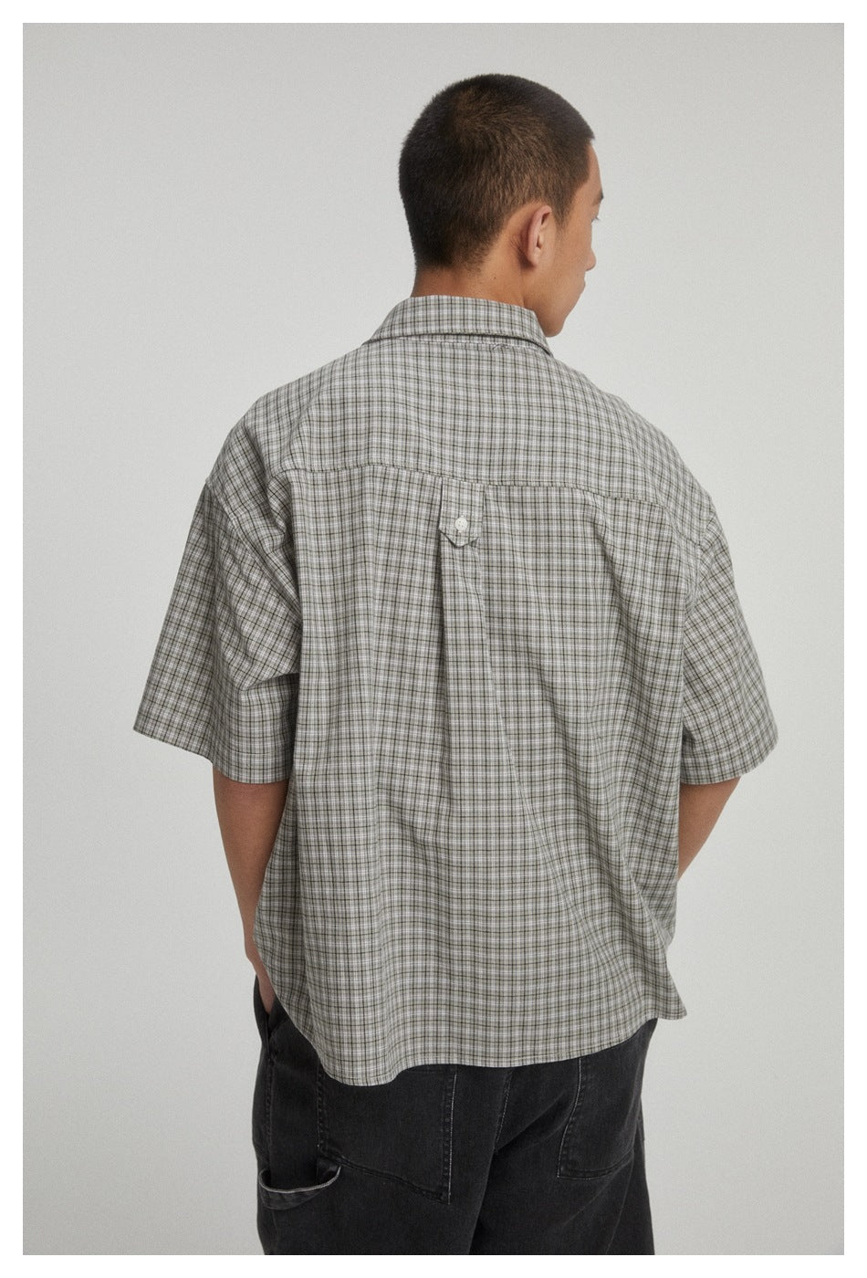 Back view of a person wearing a loose gray plaid short-sleeve button-up shirt.