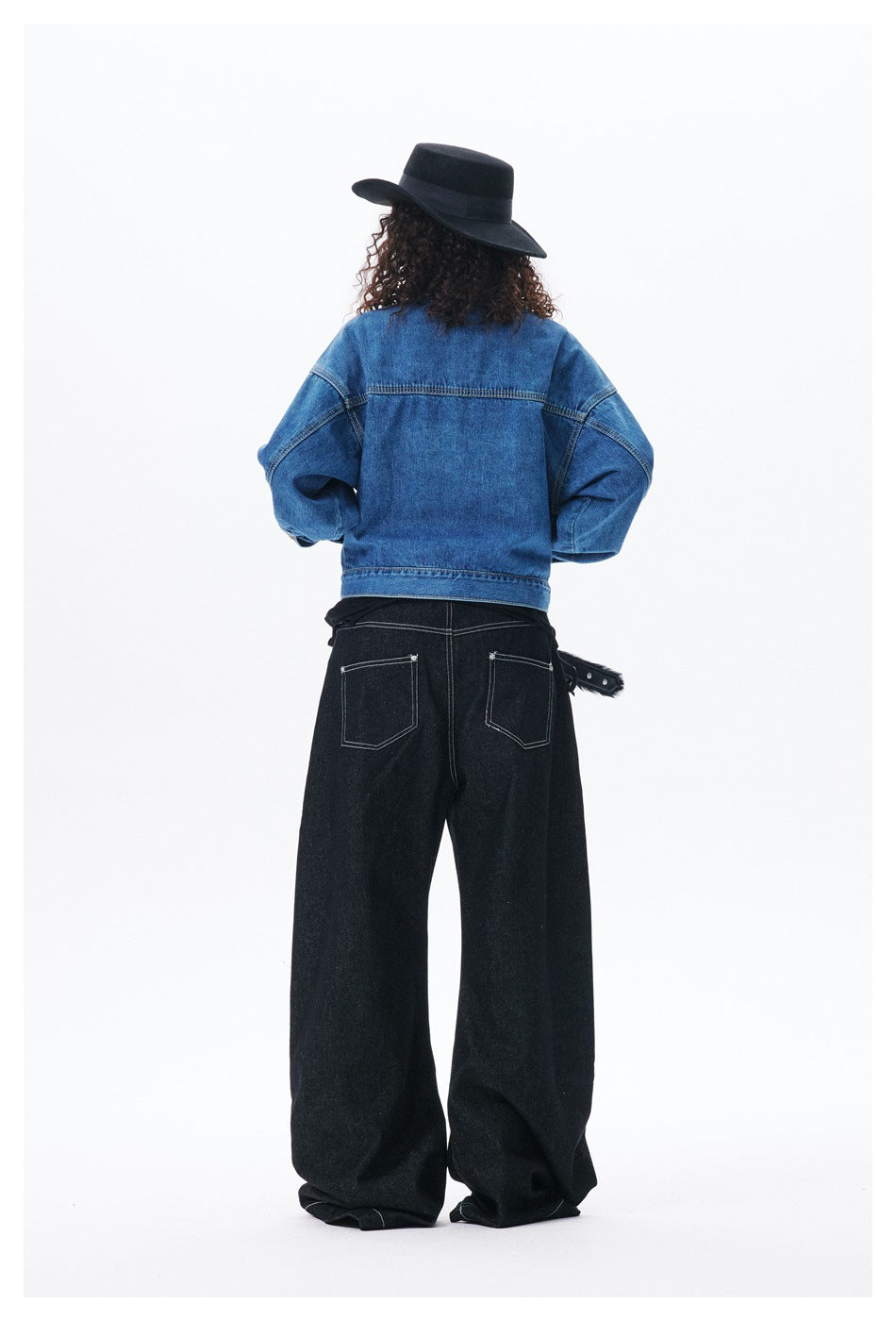 Back view of a model wearing a blue denim jacket and loose black jeans with outlined back pockets.