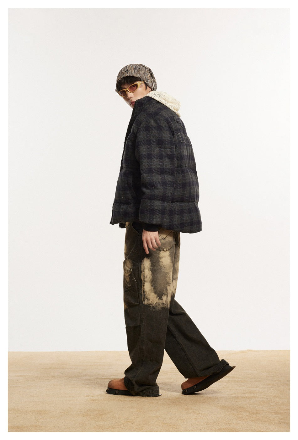Model in side view wearing the plaid puffer jacket with stand collar, beanie, tinted sunglasses and wide dark pants