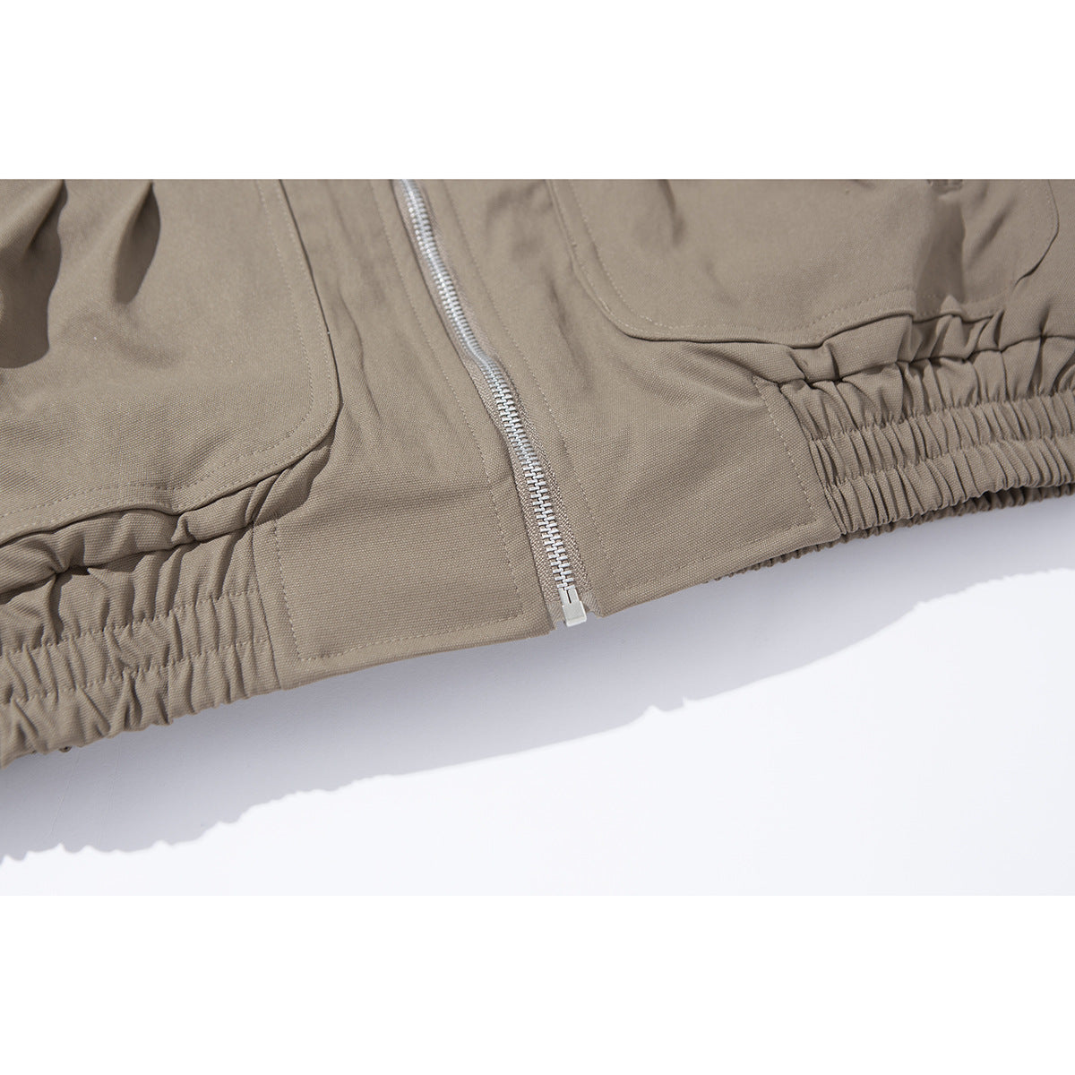 Close-up of a taupe jacket’s front zipper, flap pocket and gathered elastic hem.
