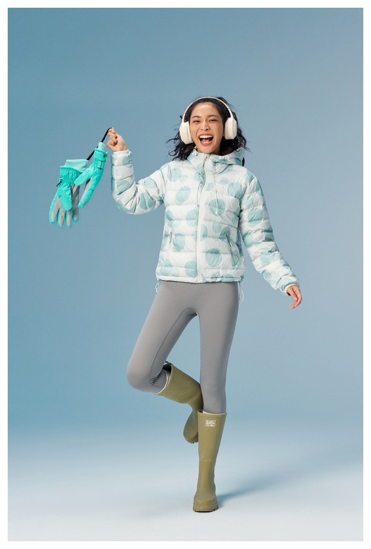 Model wearing a white quilted hooded jacket with large aqua circular prints, gray leggings, and olive rain boots.