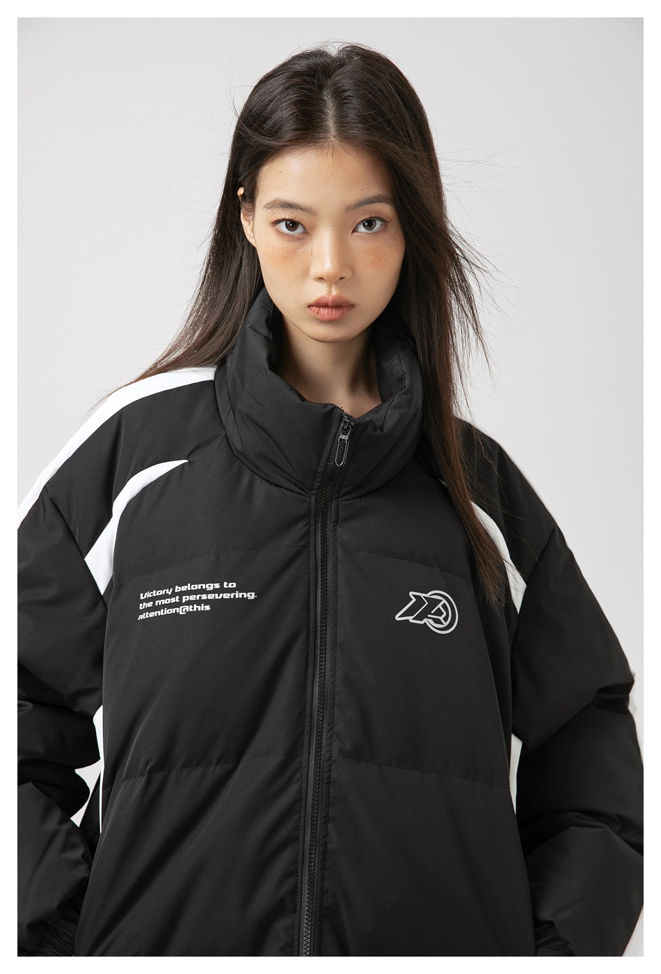 Close view of a model wearing a black padded jacket, showing its high collar, zipper, white stripes, and chest graphics.