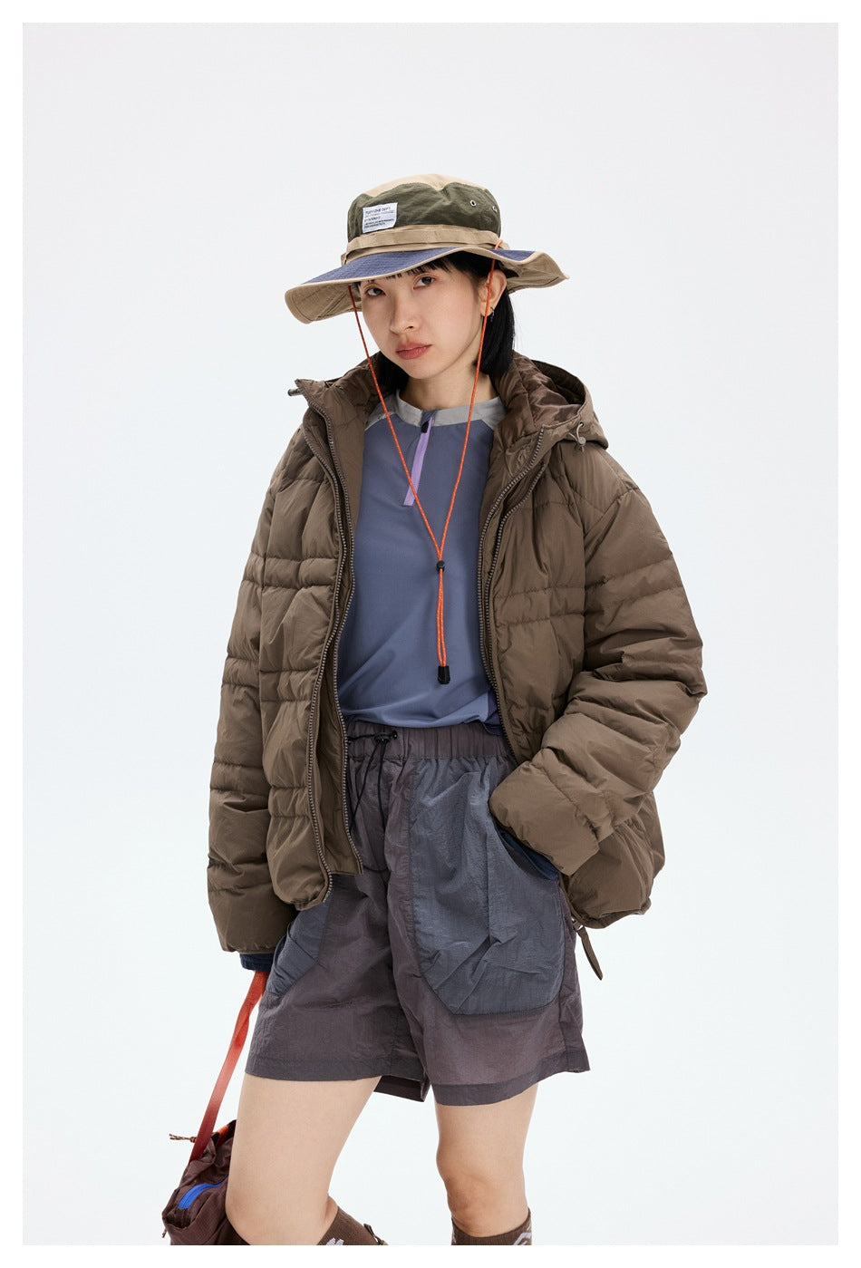 Model wearing an open brown quilted puffer jacket over a blue top and gray shorts, with a wide-brim hat.