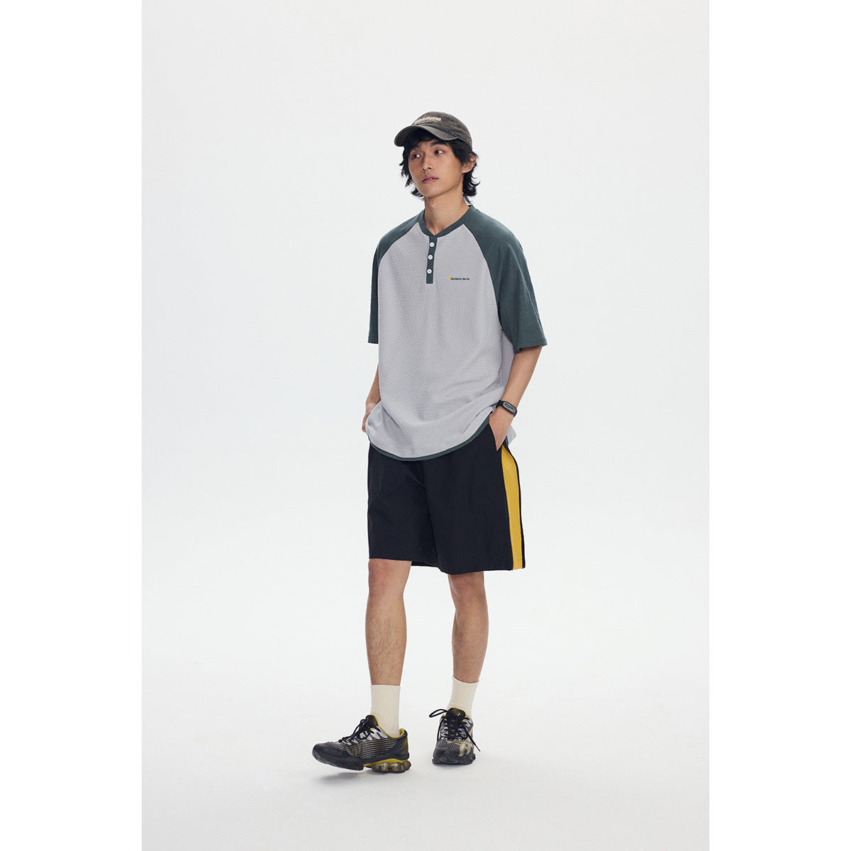 Full-length view of a model wearing a gray and dark green raglan henley with black shorts featuring a yellow side stripe.