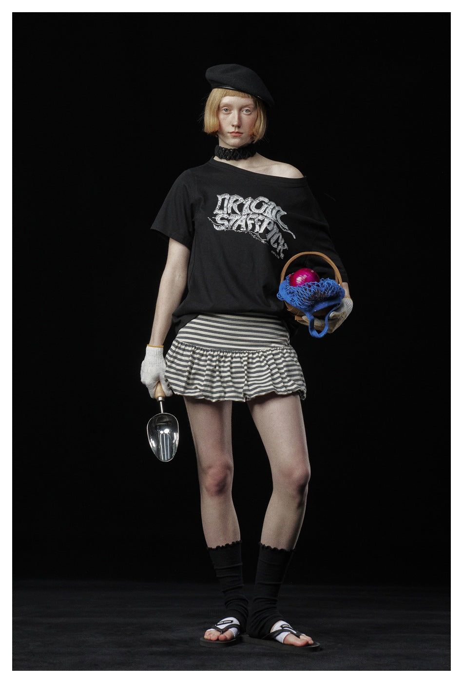 Model wearing black off-shoulder-style graphic T-shirt with striped mini skirt, beret and sandals against a black backdrop