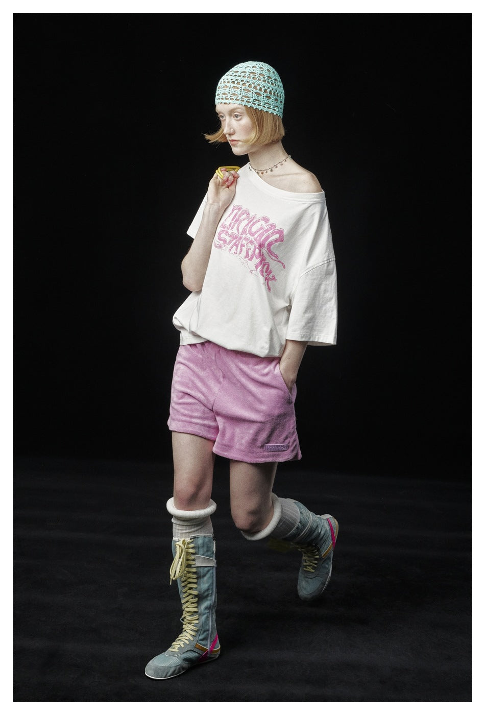 Model wearing an oversized white off-shoulder graphic T-shirt, lilac shorts, and a mint crocheted cap against a black