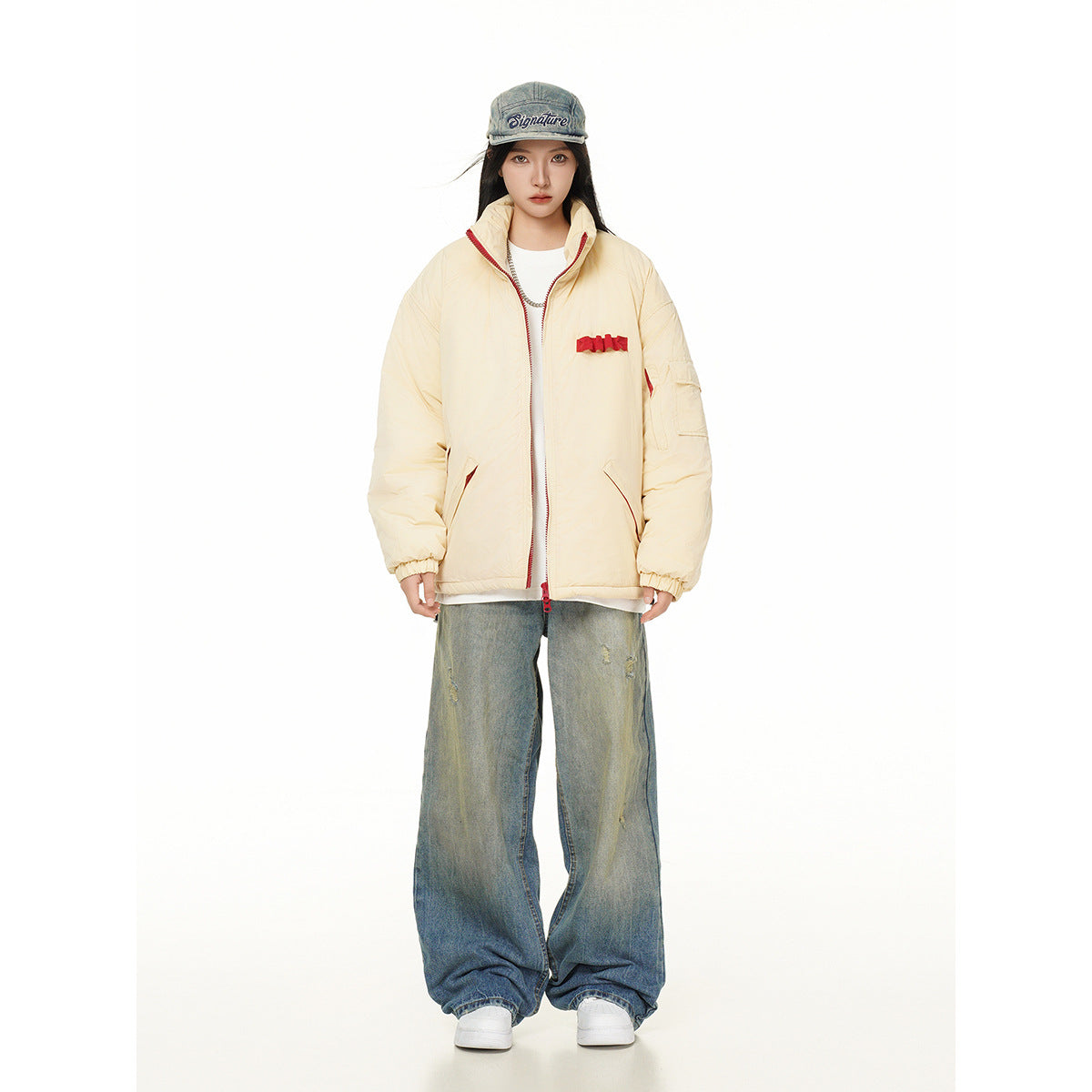 Model wearing a cream padded jacket with red trim and chest accents, wide distressed jeans, and a denim cap.