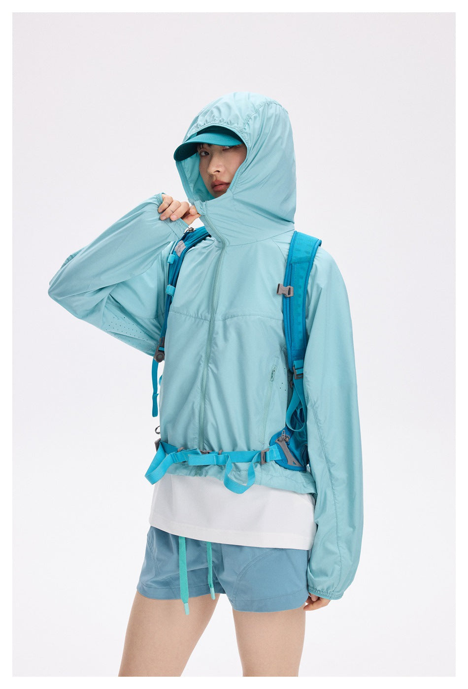 Model wears a pale aqua jacket with its hood up over a teal cap, shown from a slight side angle.