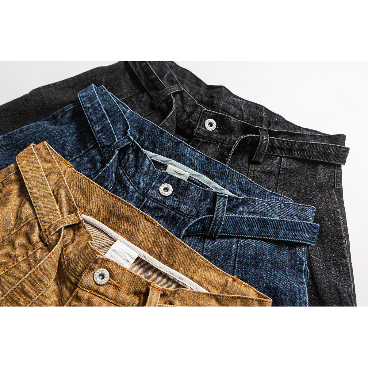 Close-up of three pairs of pleated trousers in ochre, blue denim, and black, arranged overlapping.