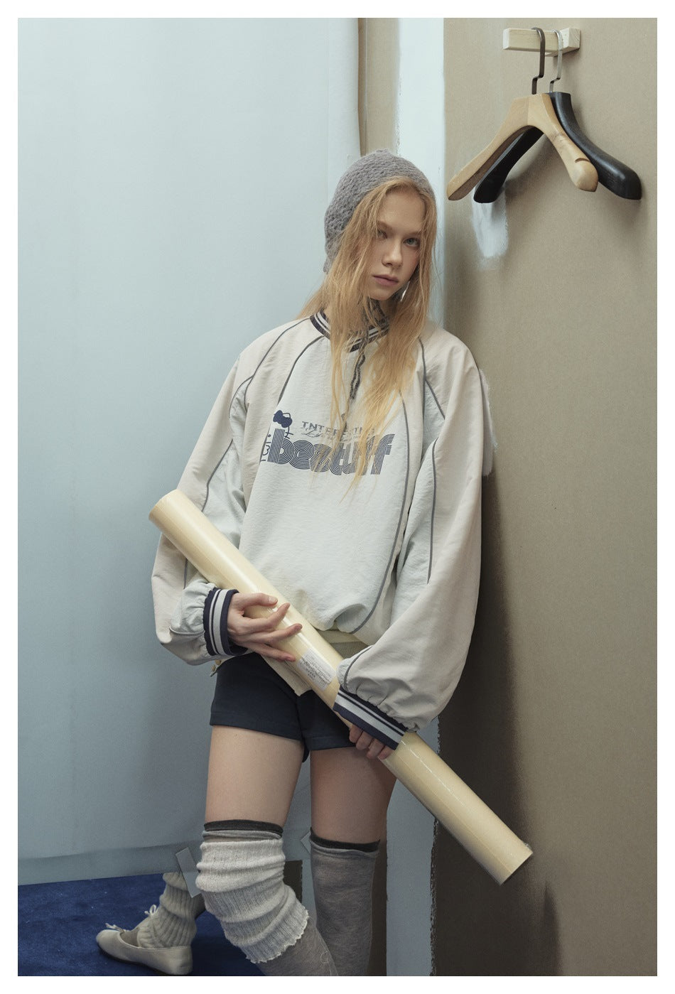 Model wearing a cream oversized graphic pullover with gray panels and striped trim, paired with dark shorts and slouchy leg