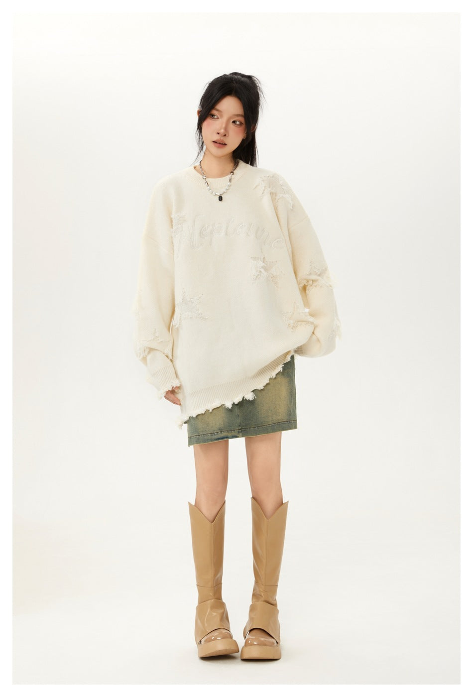 Model wearing an oversized ivory distressed sweater with tonal chest lettering, a faded denim skirt and tan boots.