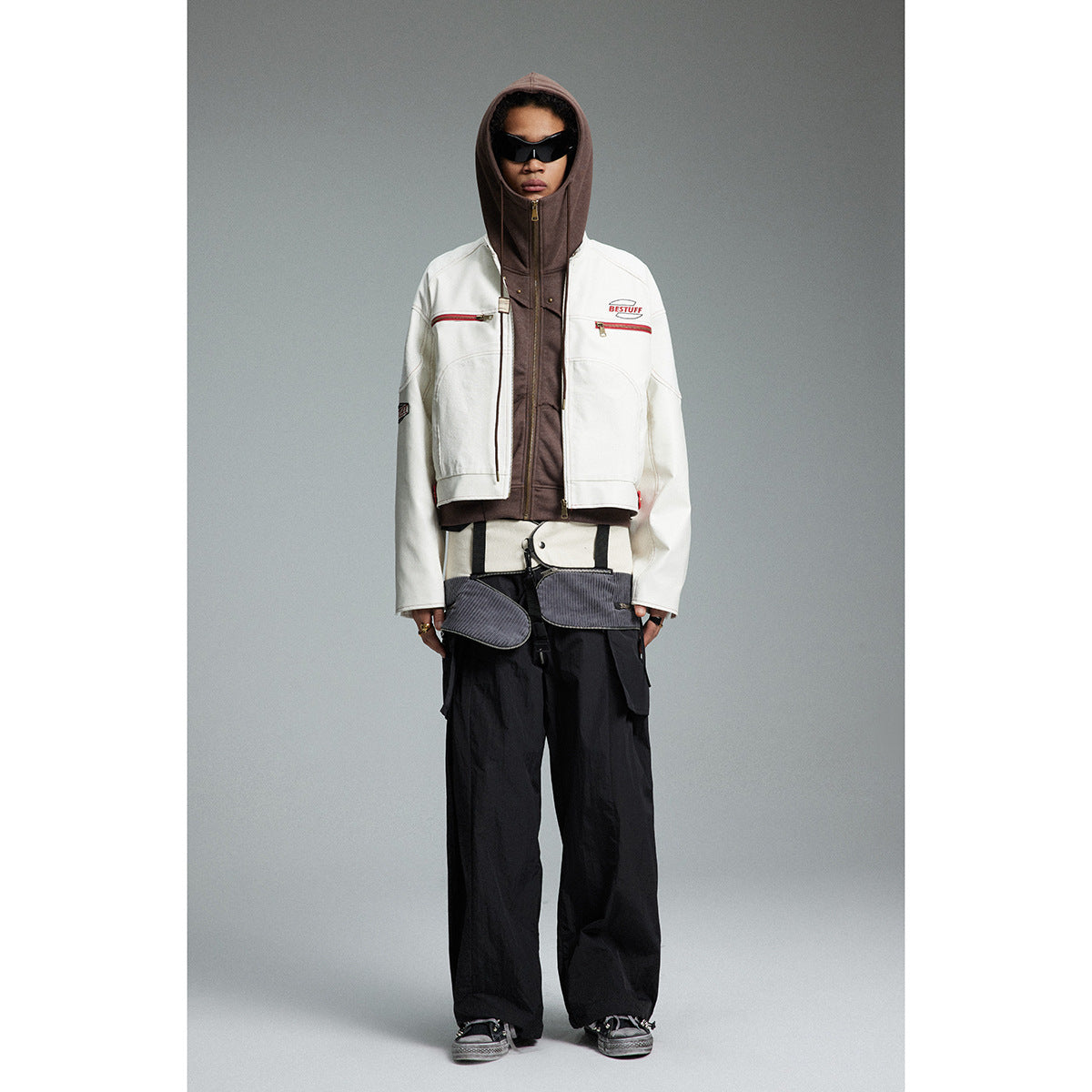 Full-length view of a model in a white jacket, zipped brown hood, black sunglasses, and wide black trousers.
