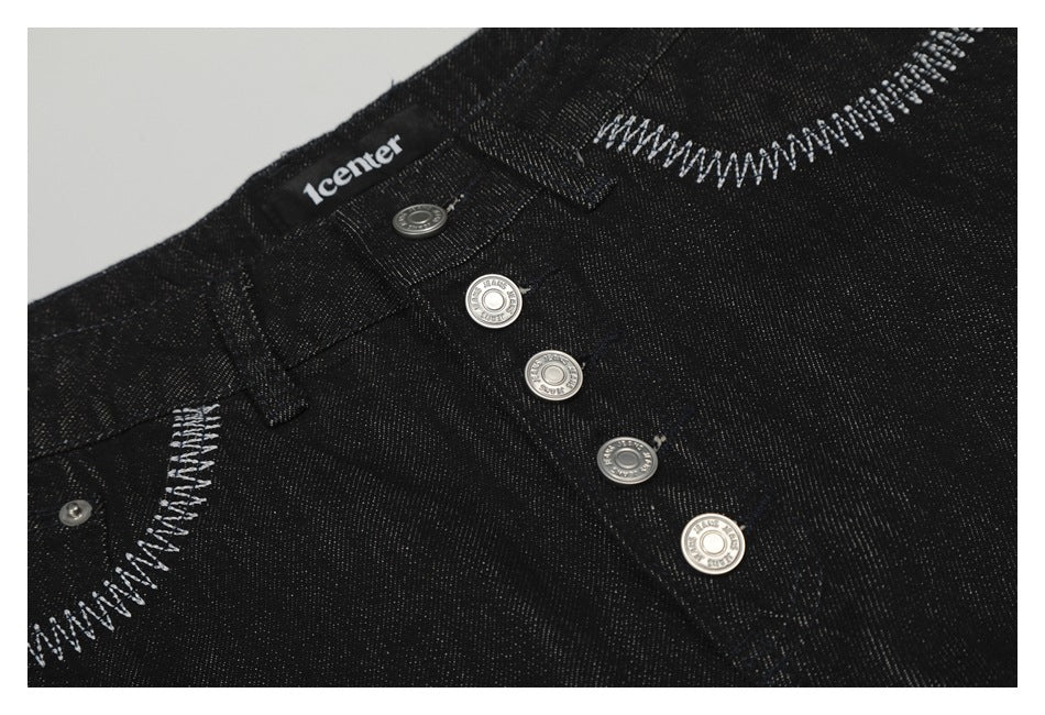 Close-up of black jeans showing the silver button fly, waistband label, and contrasting white pocket stitching.