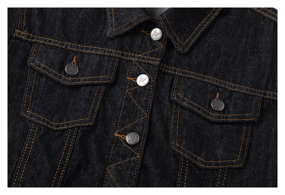 Close-up of a black denim jacket front with two flap pockets, metal buttons and orange stitching.