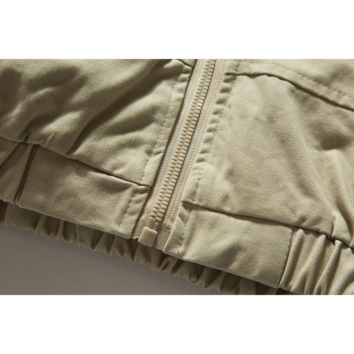 Close-up of a light khaki jacket's front zipper, stitched panels, and gathered hem.