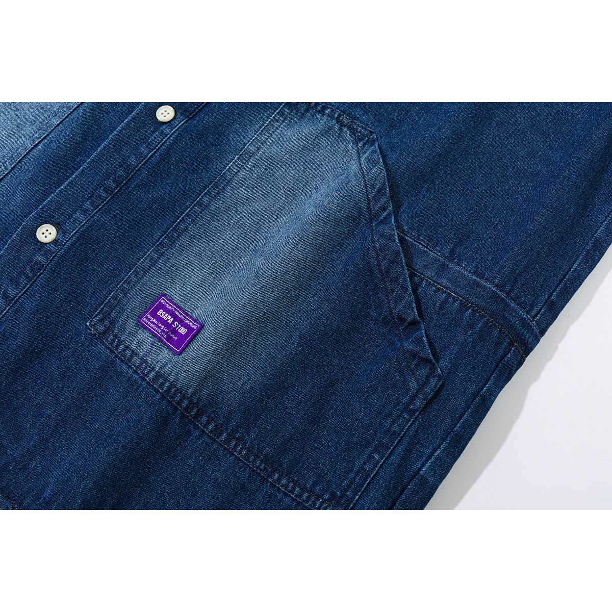 Close-up of a blue denim shirt front pocket, placket and white buttons, with a small purple label.