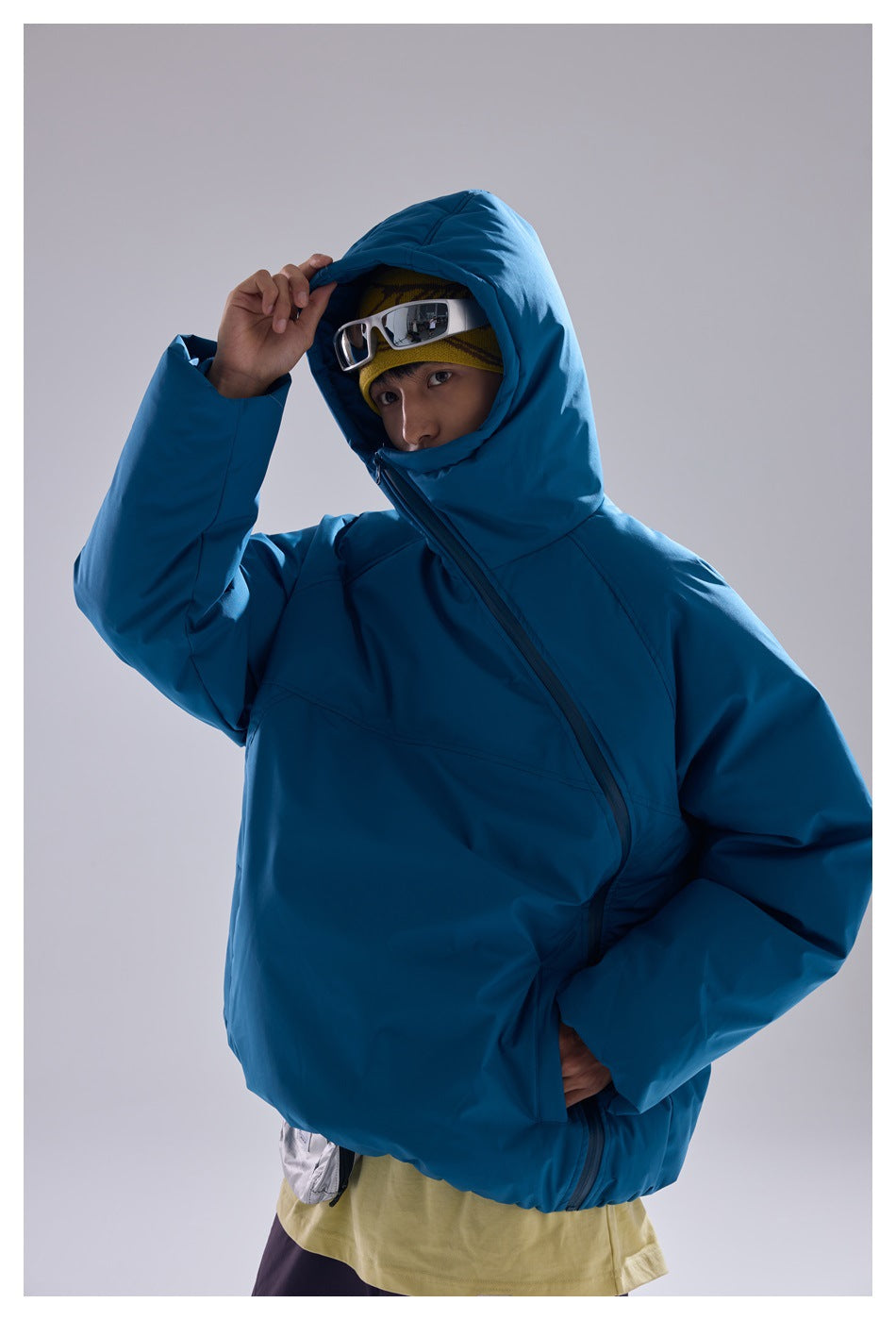 Person wearing a blue hooded padded jacket with diagonal zipper, hand raised to the hood, goggles on a yellow beanie