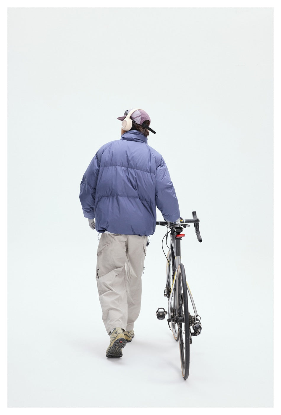 Rear view of a person in a blue puffer jacket and light cargo pants walking beside a road bicycle.