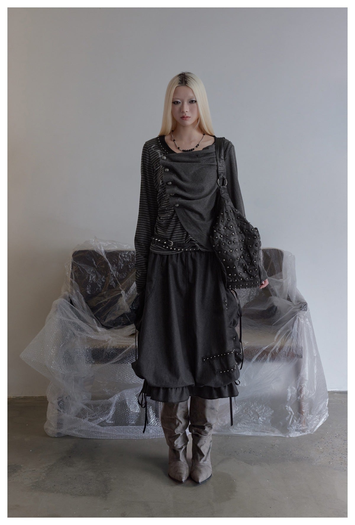 Model standing in a black gathered skirt with studded trim and ribbon ties, worn with a striped top and gray boots