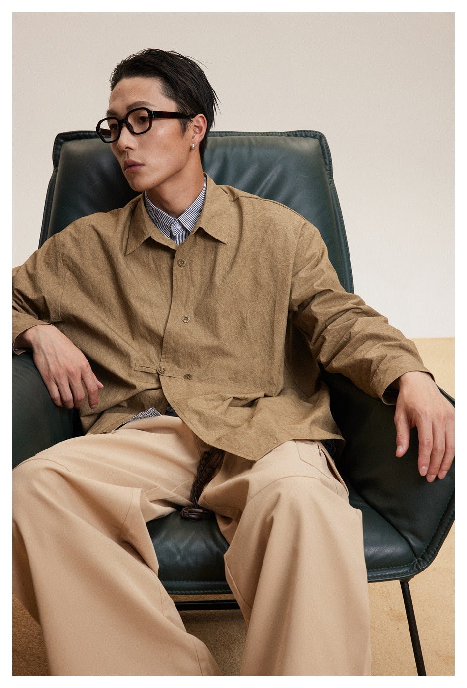 Seated model wearing a loose brown button-up shirt with a pointed collar and beige trousers.