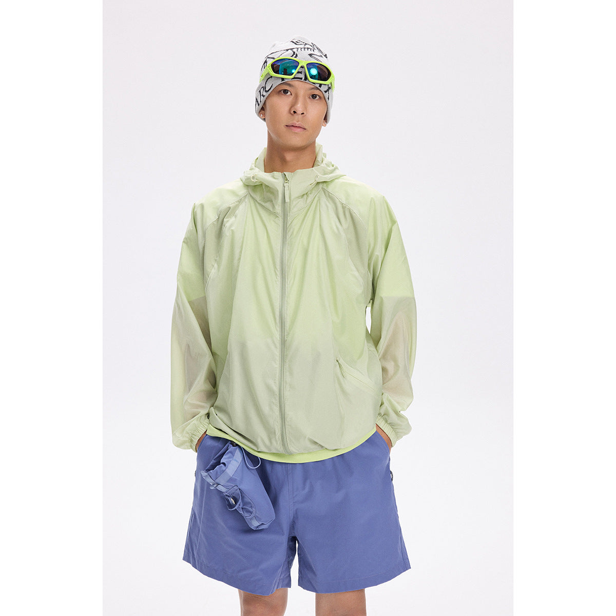 Model wearing a pale green hooded zip jacket and blue shorts, with a patterned beanie and bright sunglasses.