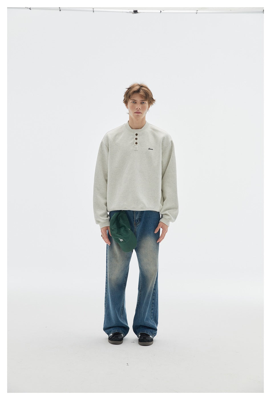 Model wearing light heather gray henley sweatshirt with wide-leg jeans, holding a green cap, front view