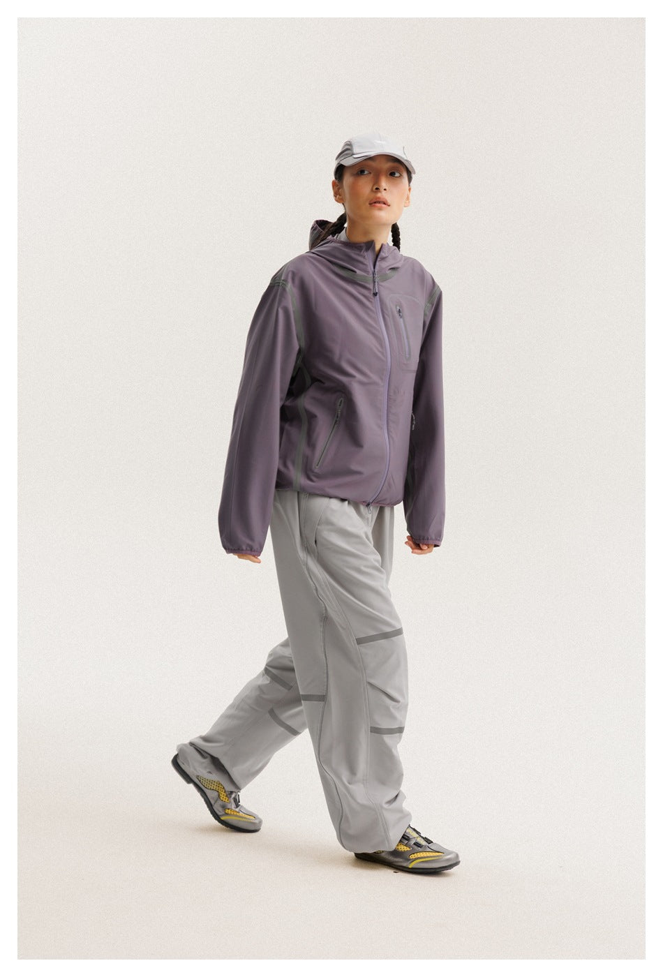 Model walking in a muted purple hooded jacket and loose gray pants with curved trim.