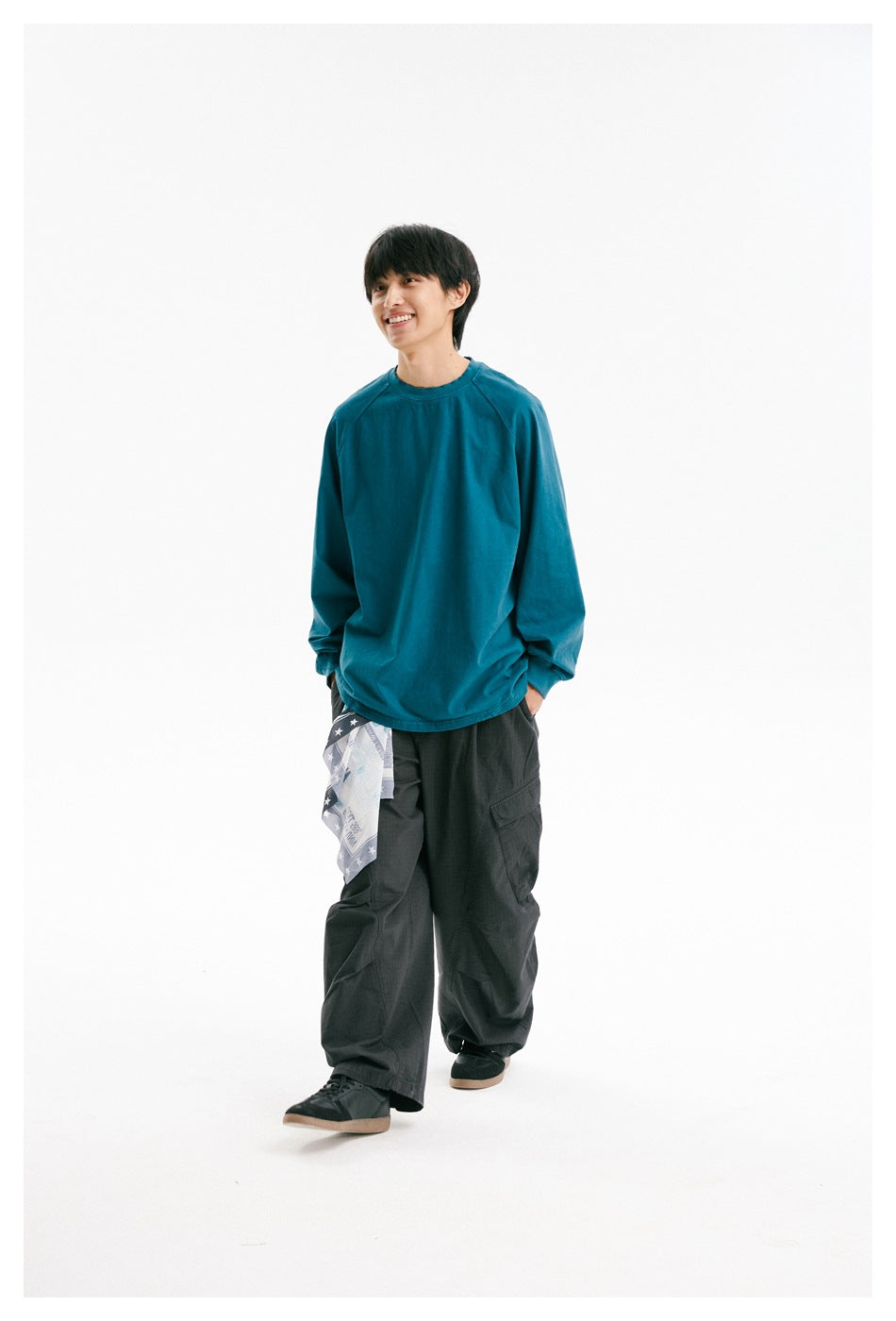 Model smiling in a teal raglan long-sleeve shirt and dark cargo trousers, walking against a white studio background.
