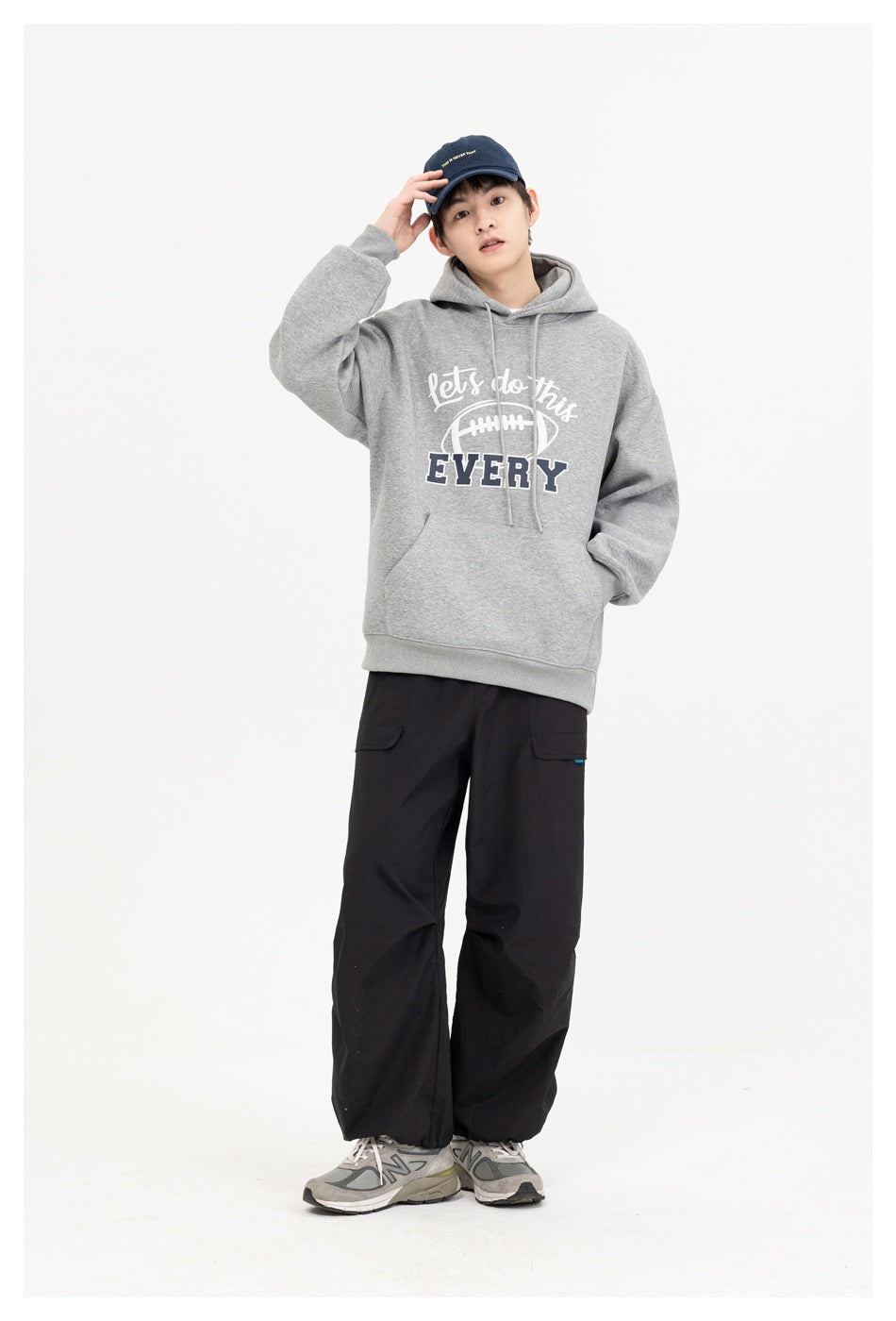 Full-length front view of a model wearing a gray football-graphic hoodie, black cargo pants, and a navy cap.