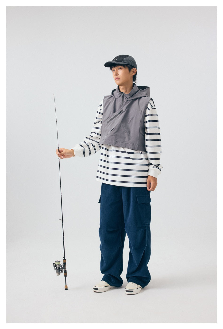 Full-length model wears a gray hooded vest over a striped shirt and navy cargo pants while holding a fishing rod.