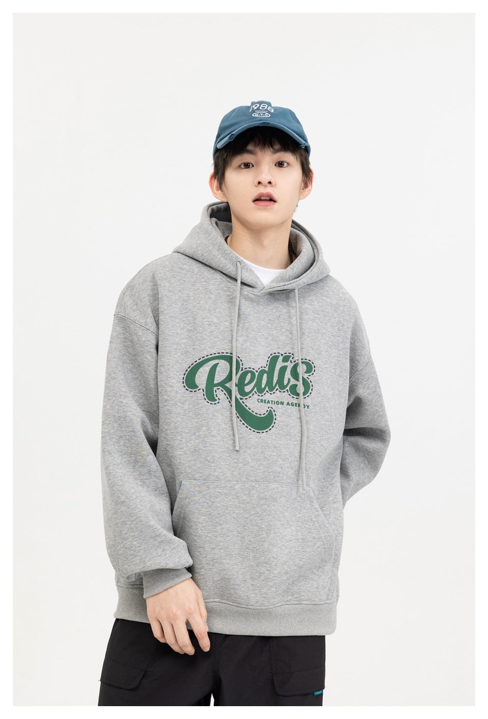 Front view of a model wearing a gray graphic hoodie with green lettering, black pants, and a blue cap.