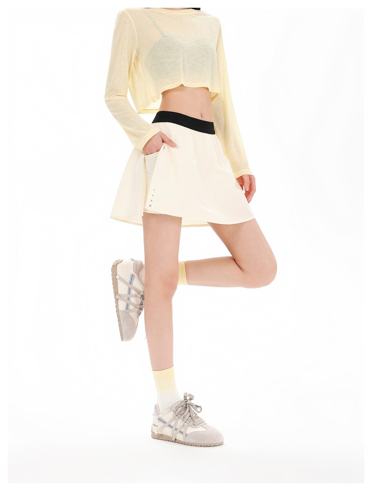 Model wearing a beige mini skirt with black waistband, hand in side pocket, with a cropped yellow top and sneakers