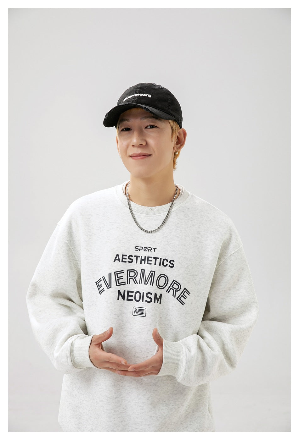 Close view of a model wearing a light gray crewneck with bold black chest lettering and a black cap.