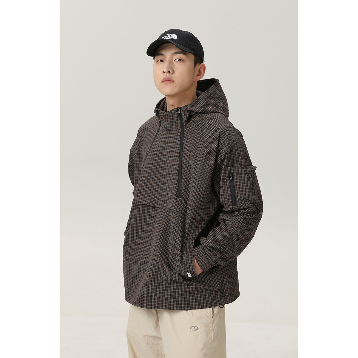 Model wearing a dark brown checked hooded pullover jacket with a front placket, kangaroo pocket and zippered sleeve pocket.