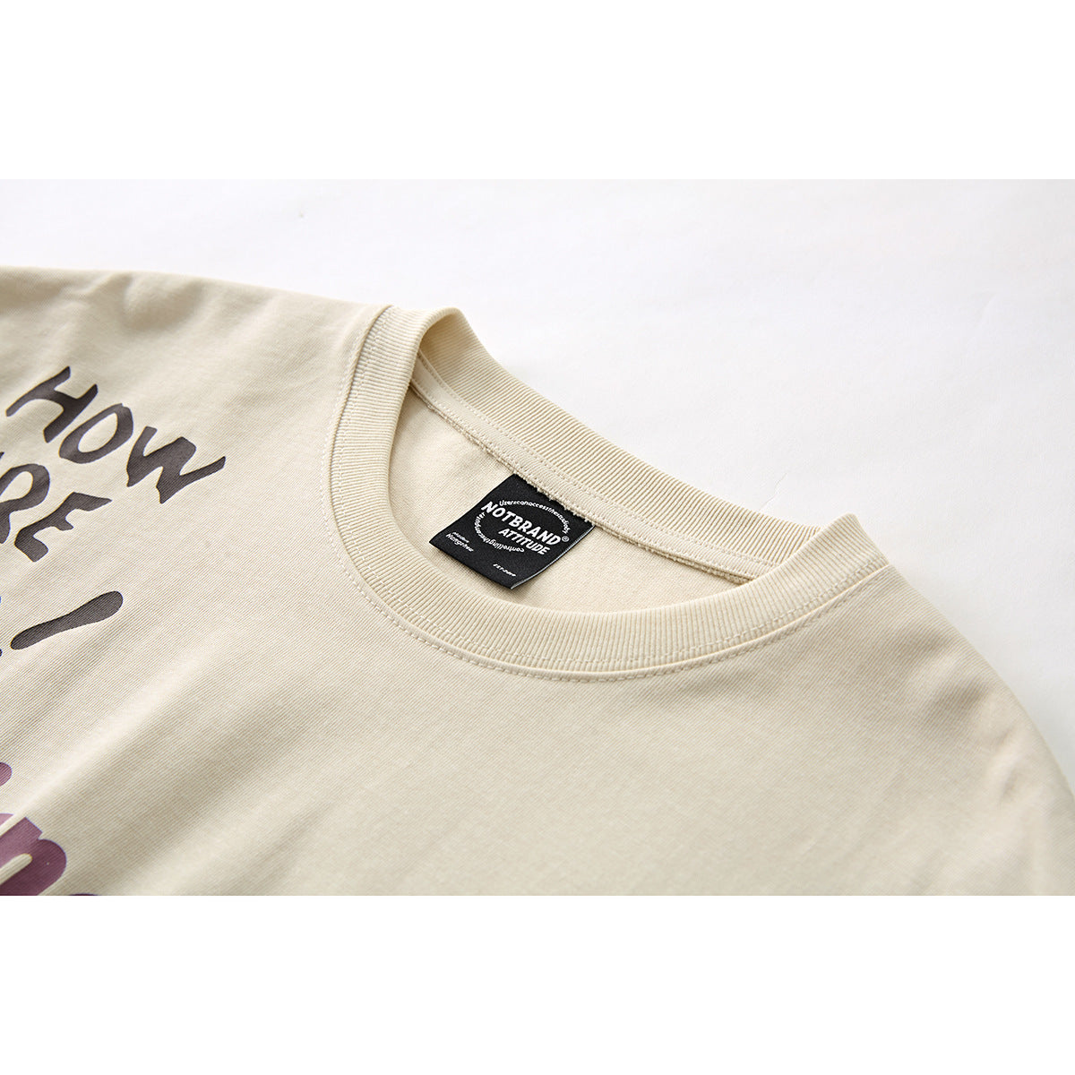 Close-up of a cream T-shirt's ribbed crew neck and black woven label, with part of the dark printed design visible.